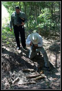 Texas Ranger taking a photo of objects on the ground in a forest with a man standing behind him